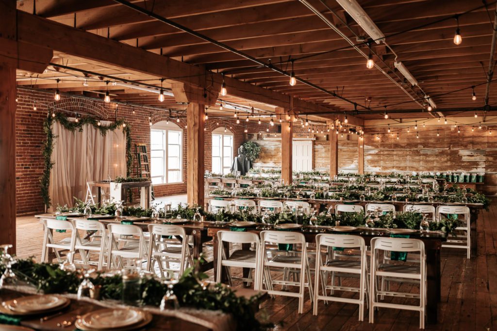 wedding reception area in a brick and mortar wedding venue. Farm house tables lined with lush greenery and string lights hanging from the ceiling.