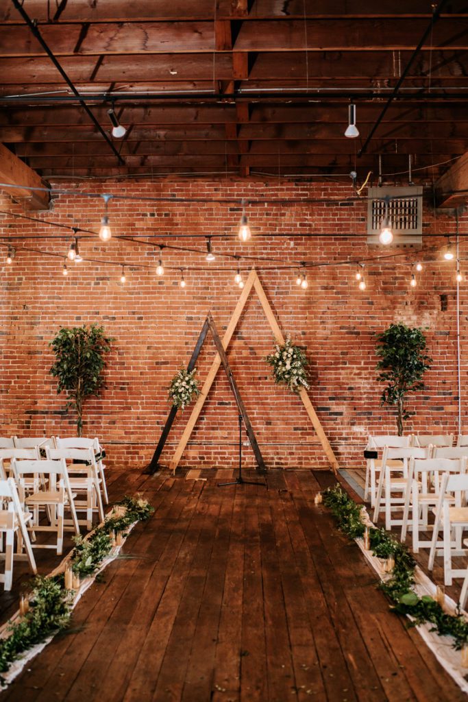 Ceremony location with two triangular wooden arbors with greenery and white flowers in a brick and mortar venue