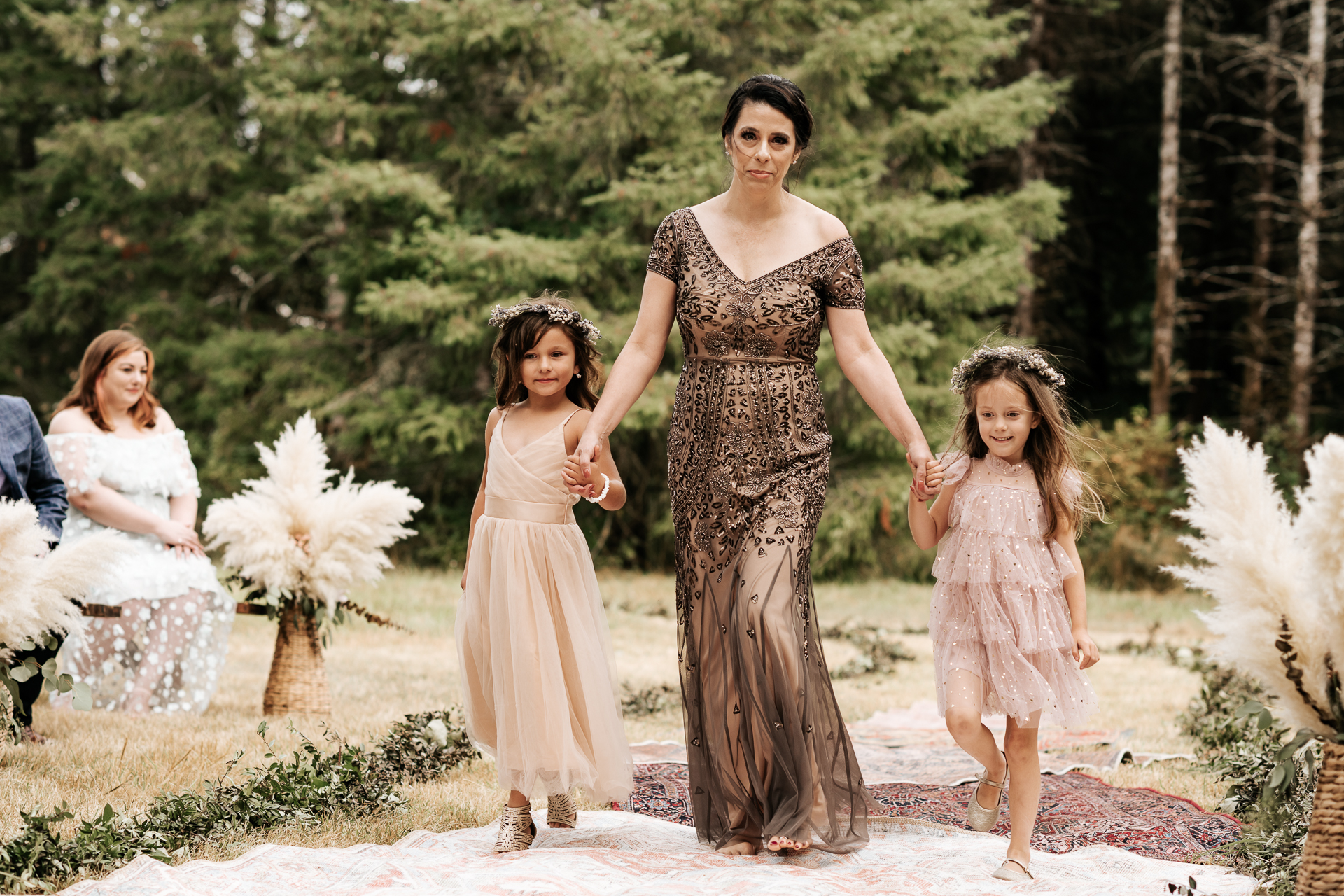 Flower girls get walked down the boho rug lined aisle by the mother of the groom