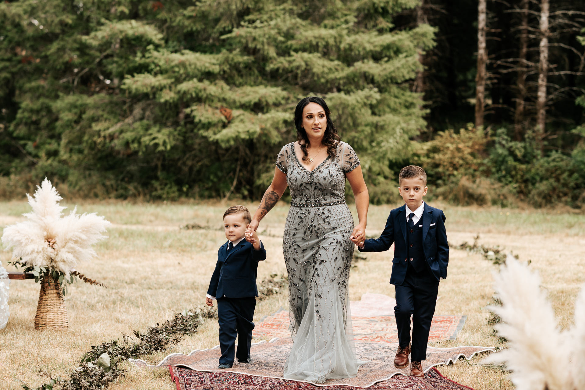 Mother of the Bride walks ring bearers down the rug lined boho aisle at a wedding