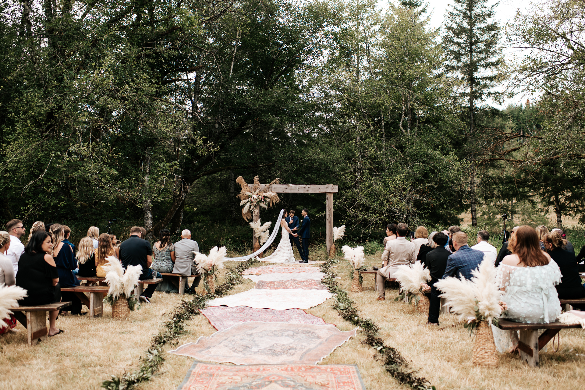 Bride and Groom standing at their pompous grass alter with a line of rugs down the aisle surrounded by friends and family