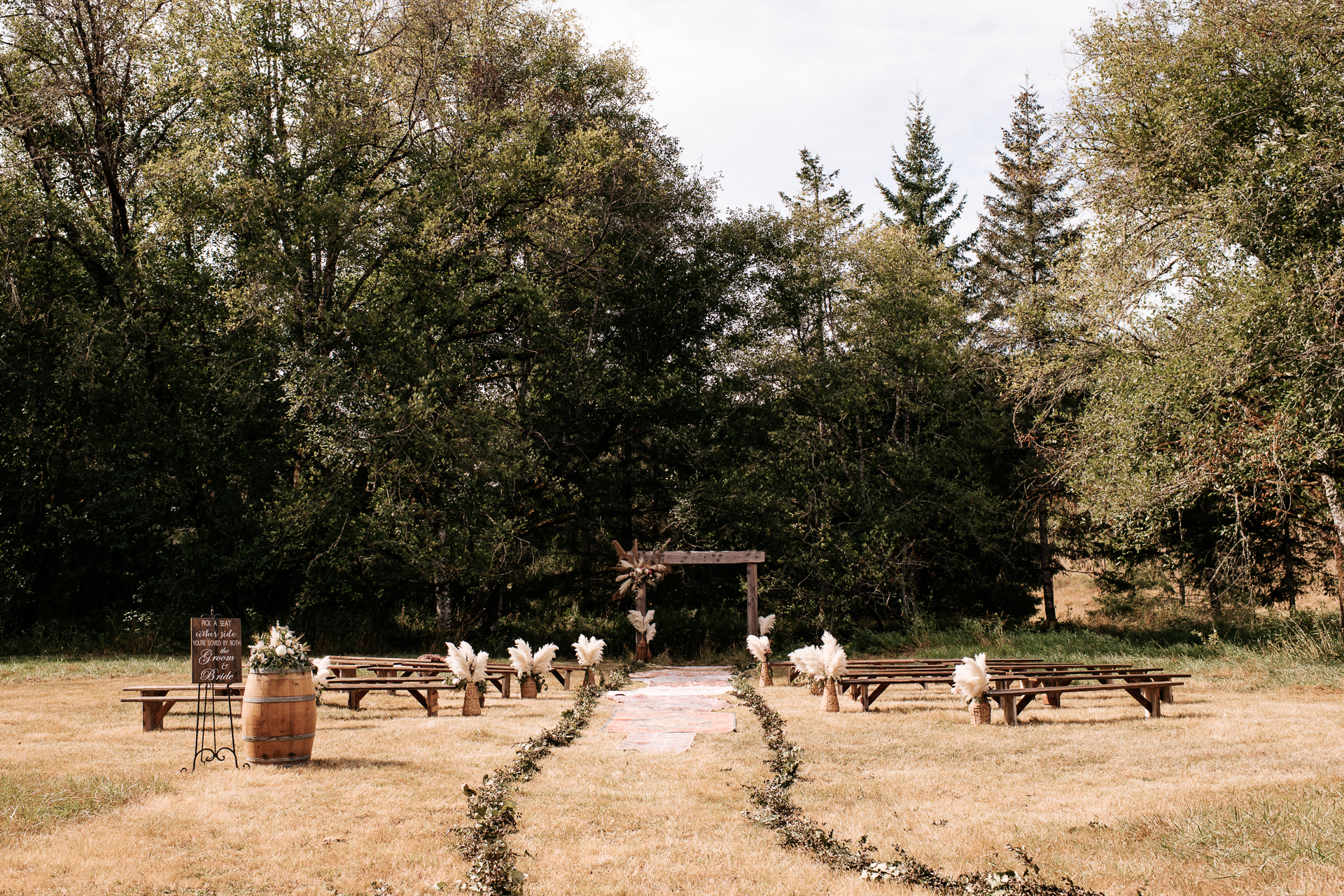 Meadow area set up for a ceremony with boho pompous grass and farm house benches to sit on with a wooden arbor and boho rugs that line the aisle