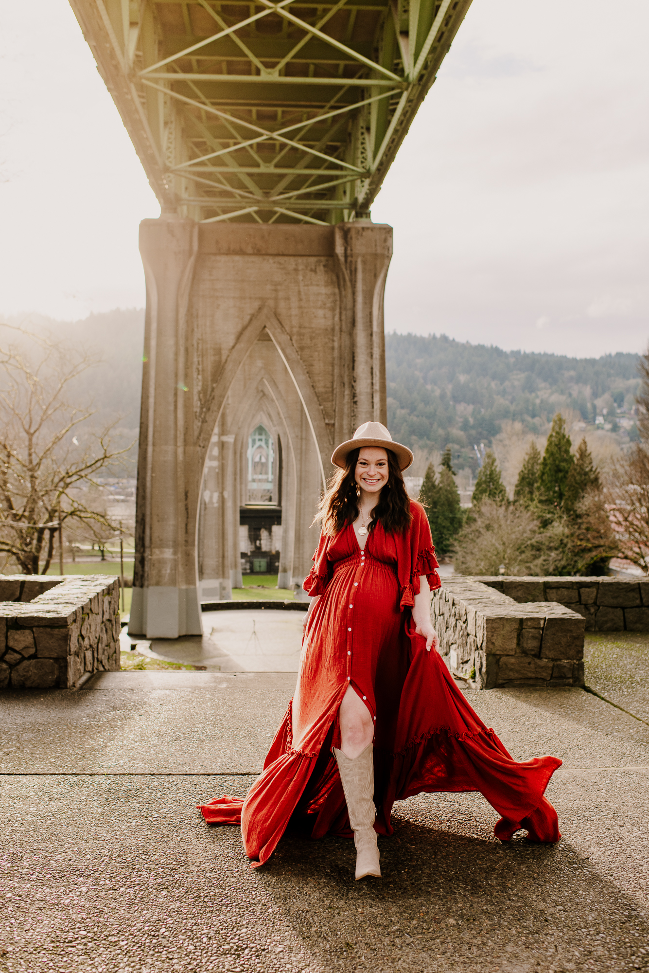 Pregnant mother walking under Cathedral Park Bridge in a red flowy maternity dress