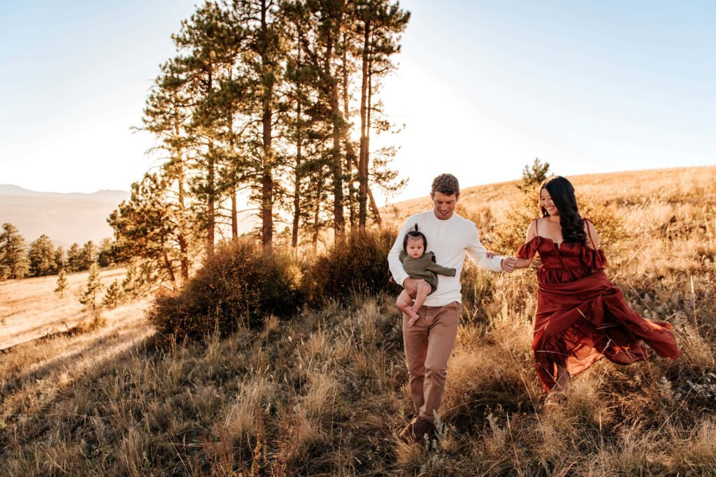 mom wearing red dress and dad wearing a off white pull over. Mom and dad holding hands walking down a grassy hill. Dad holding baby in arms.
